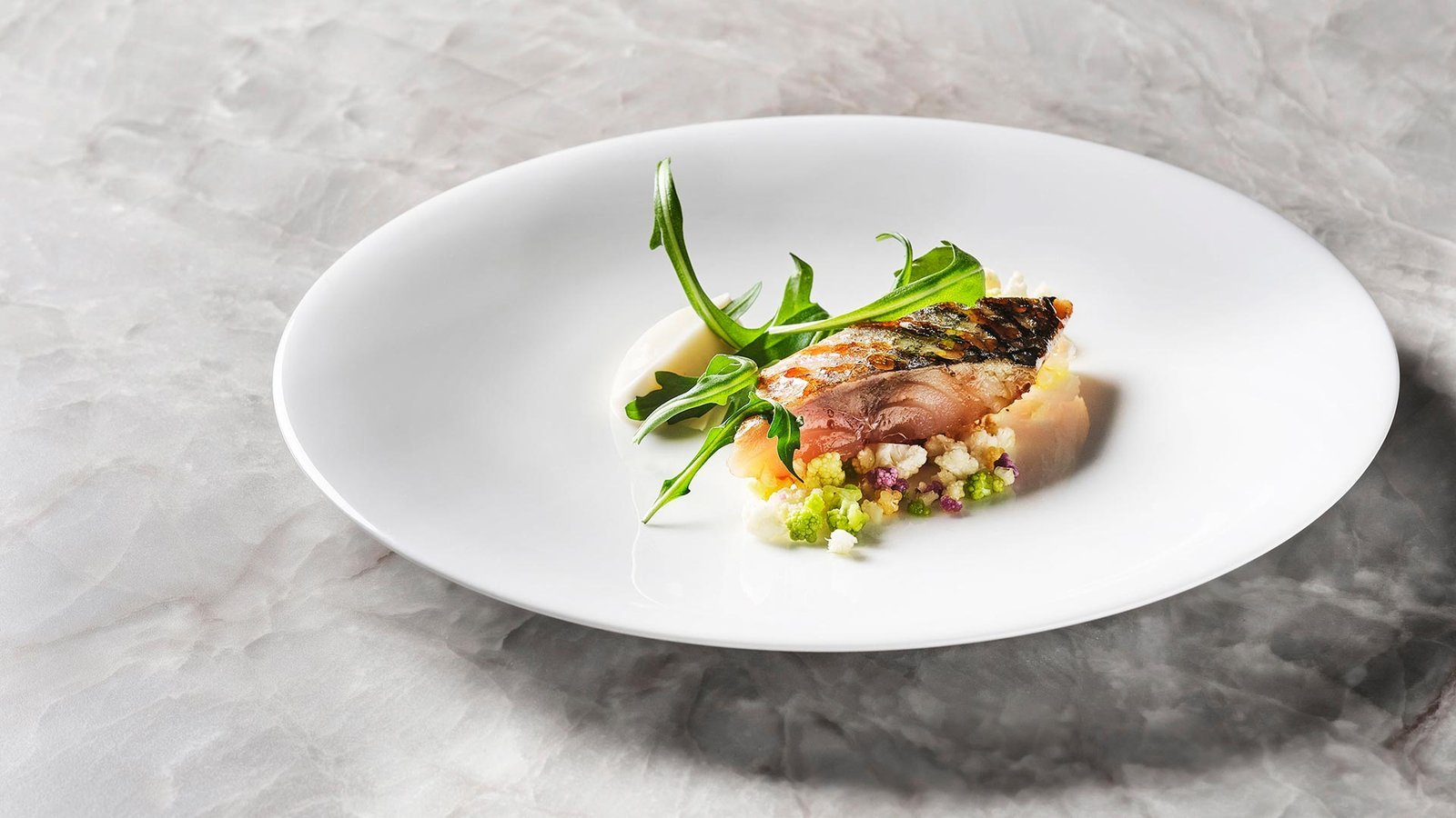 Pescado a la brasa con romanesco y rúcula sobre plato blanco y mármol.