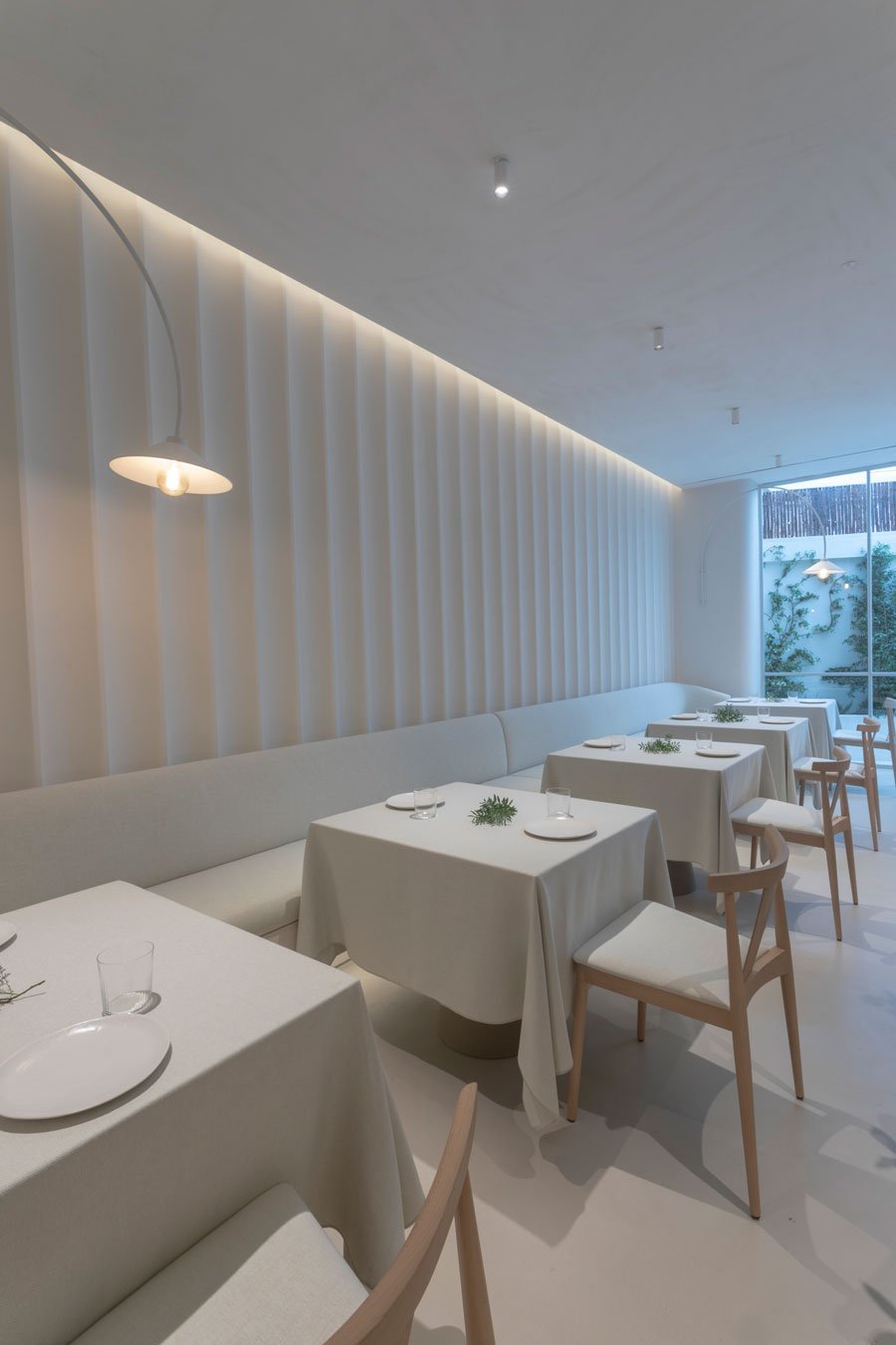 Rincón de la sala de VelascoAbellà con luz cálida y vegetación tras la cristalera.